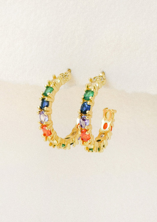 A pair of gold hoop earrings adorned with various colored gems hanging from a white paper.