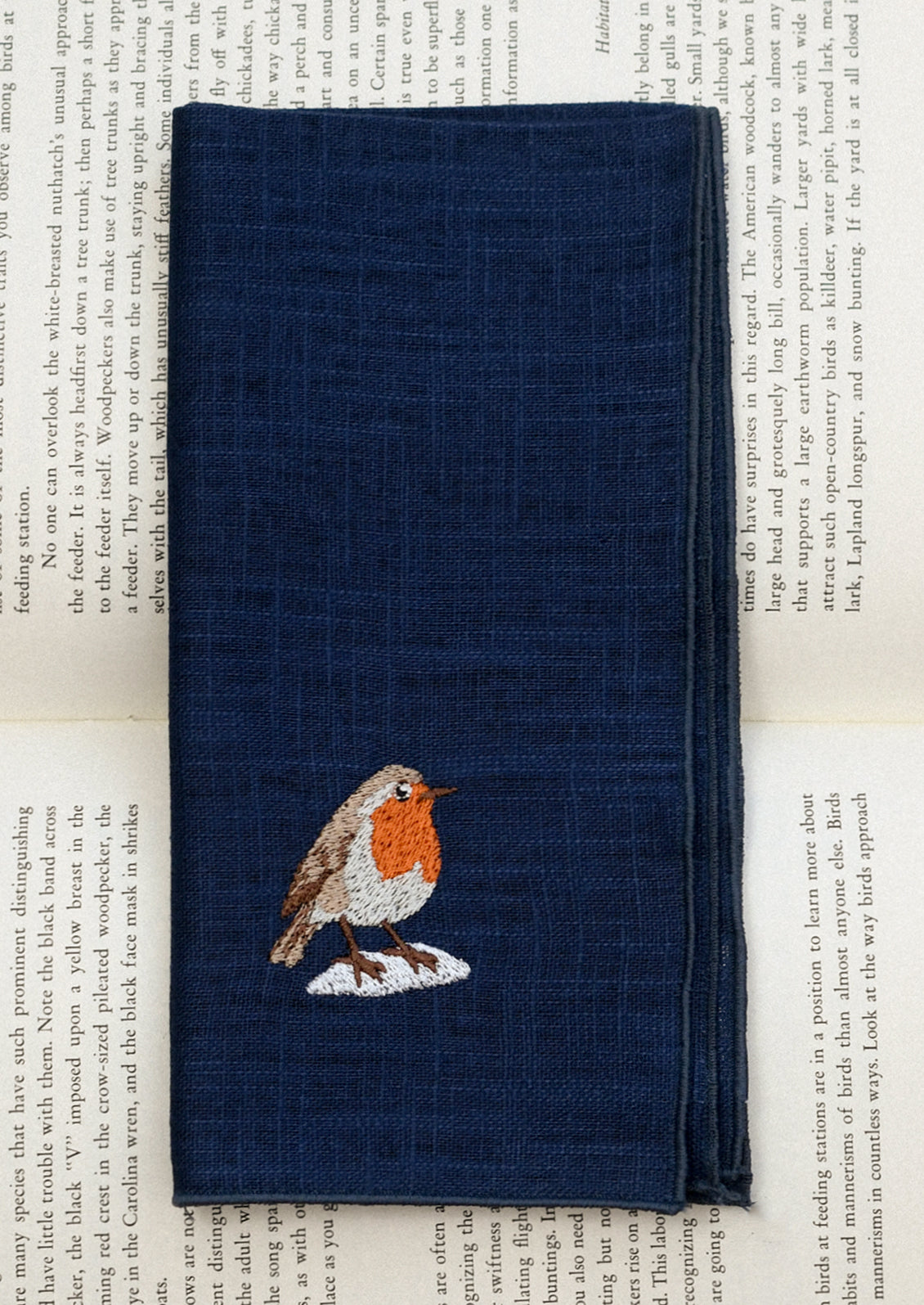 A navy blue linen napkin with tonal serged edges, and an embroidered robin at corner.