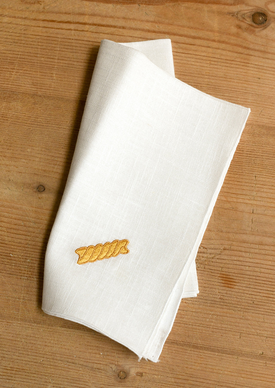 A folded white linen napkin with a small yellow embroidered pasta shape, displayed on a light brown wooden surface.