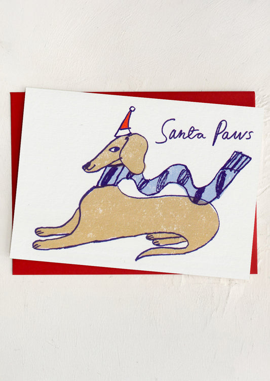 A greeting card with illustration of a brown dog wearing a Santa hat, text reads "Santa Paws"