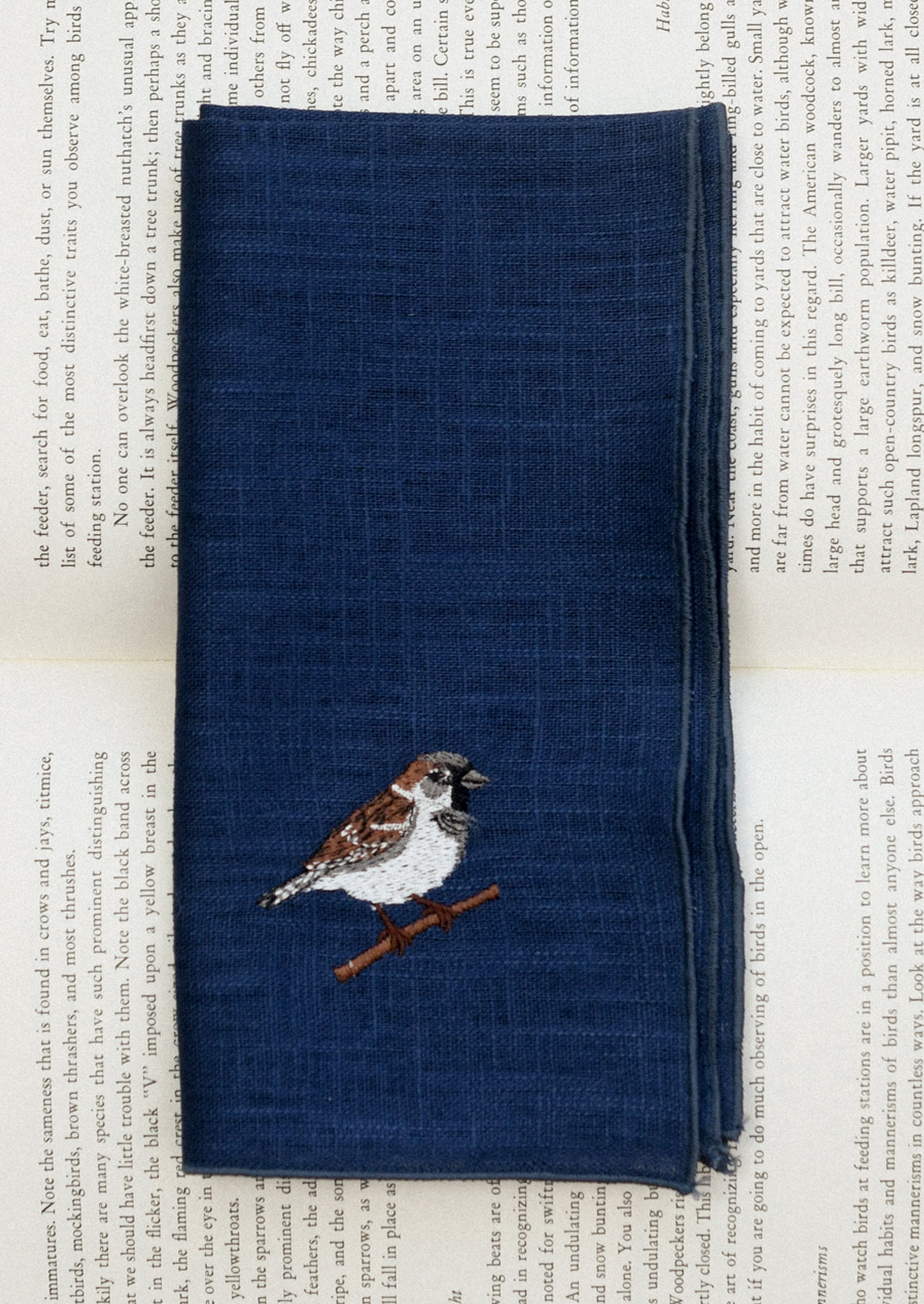 A navy blue linen napkin with tonal serged edges, and an embroidered sparrow at corner.