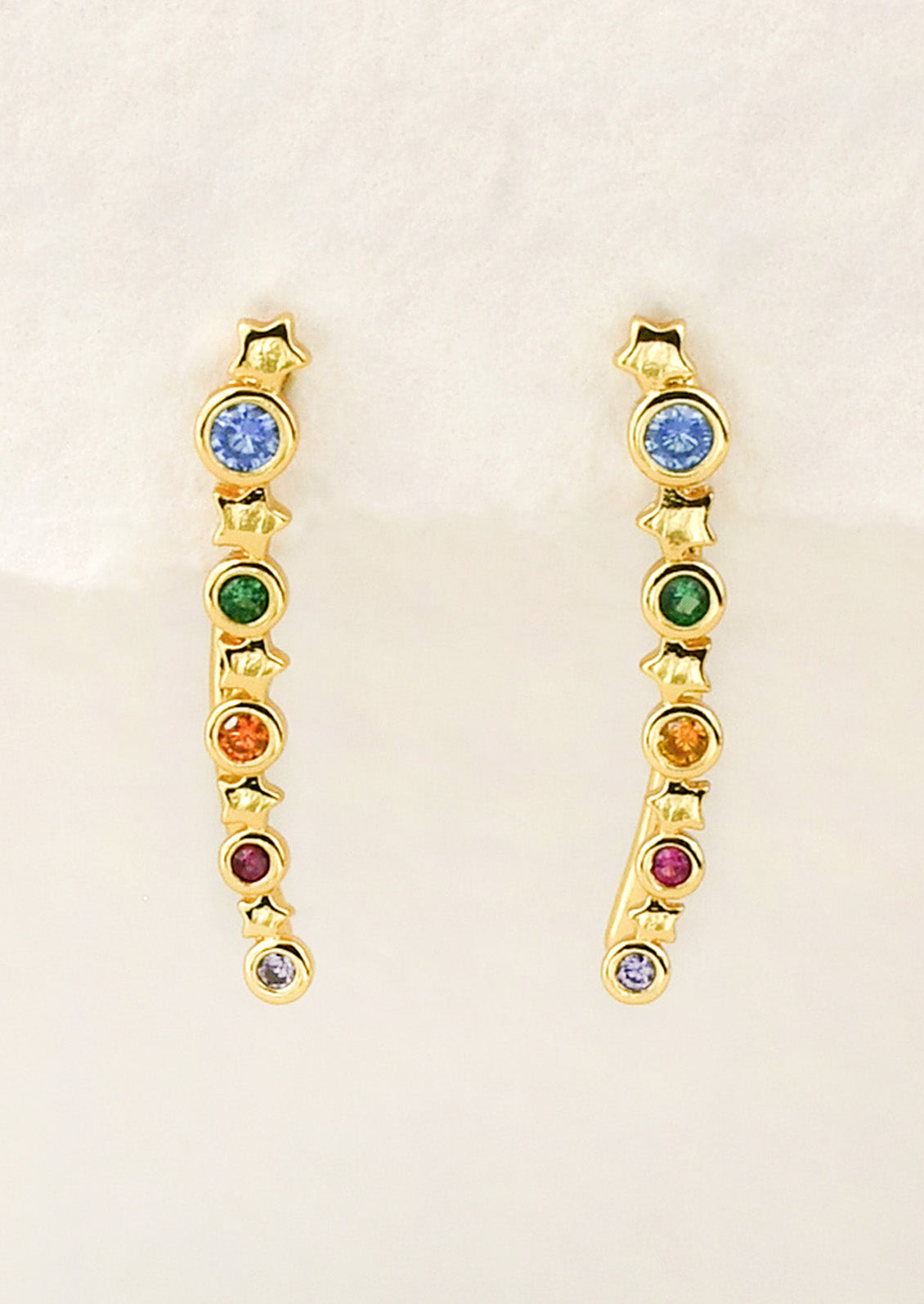 A pair of long gold earrings with a star design and colored gems throughout, hanging from a white paper.