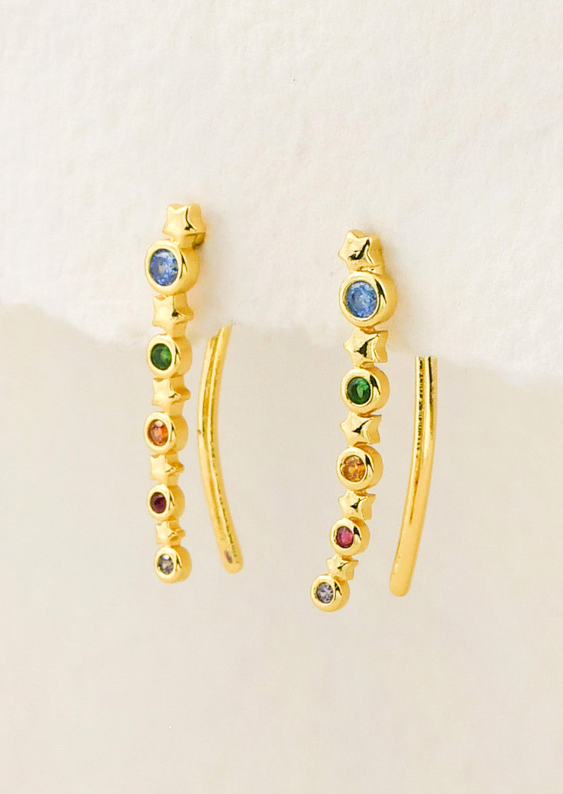 A pair of long gold earrings with a star design and colored gems throughout, hanging from a white paper.
