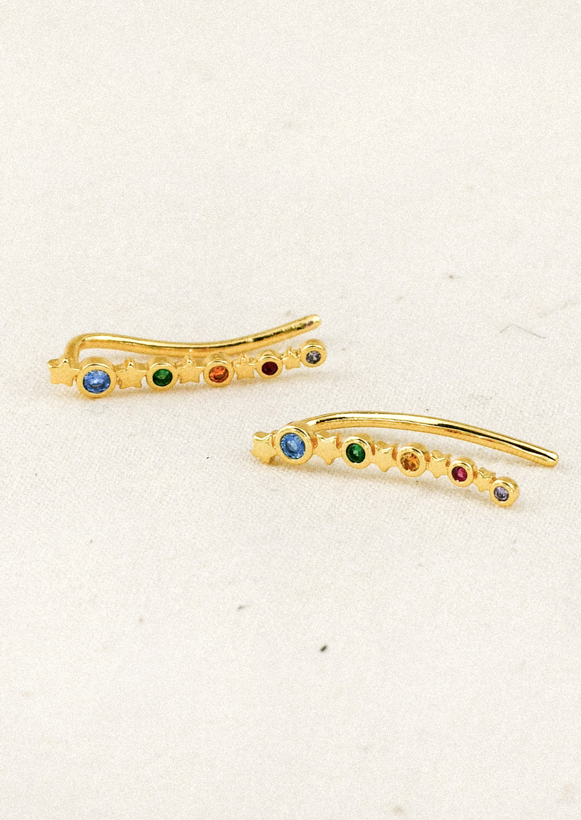 A pair of long gold earrings with a star design and colored gems throughout on a white background.