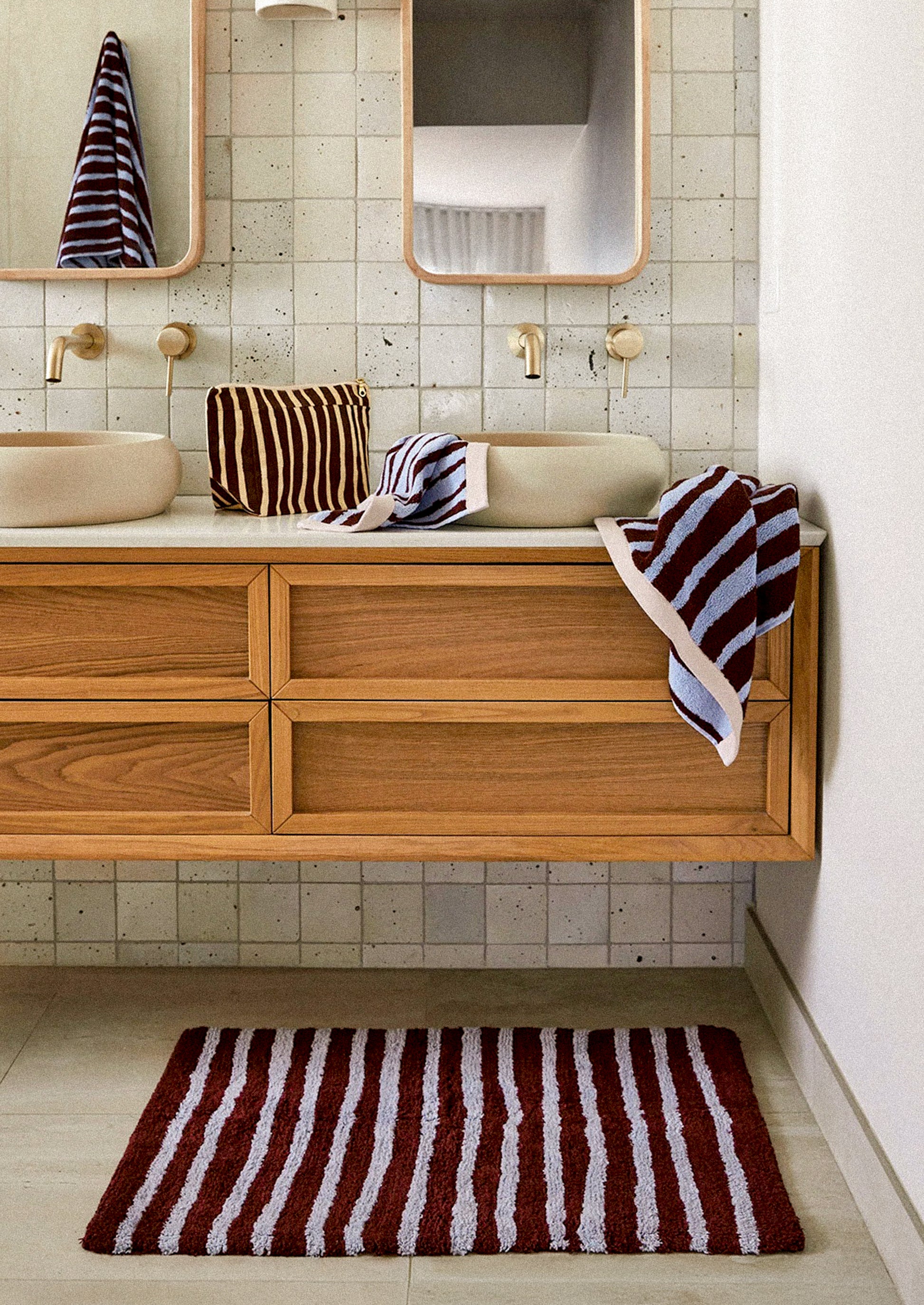 A bathroom vanity scene with a wooden floating cabinet, two cream vessel sinks, mirrors, and striped towels in dark red, white, and navy. A matching striped bath mat is on the floor.