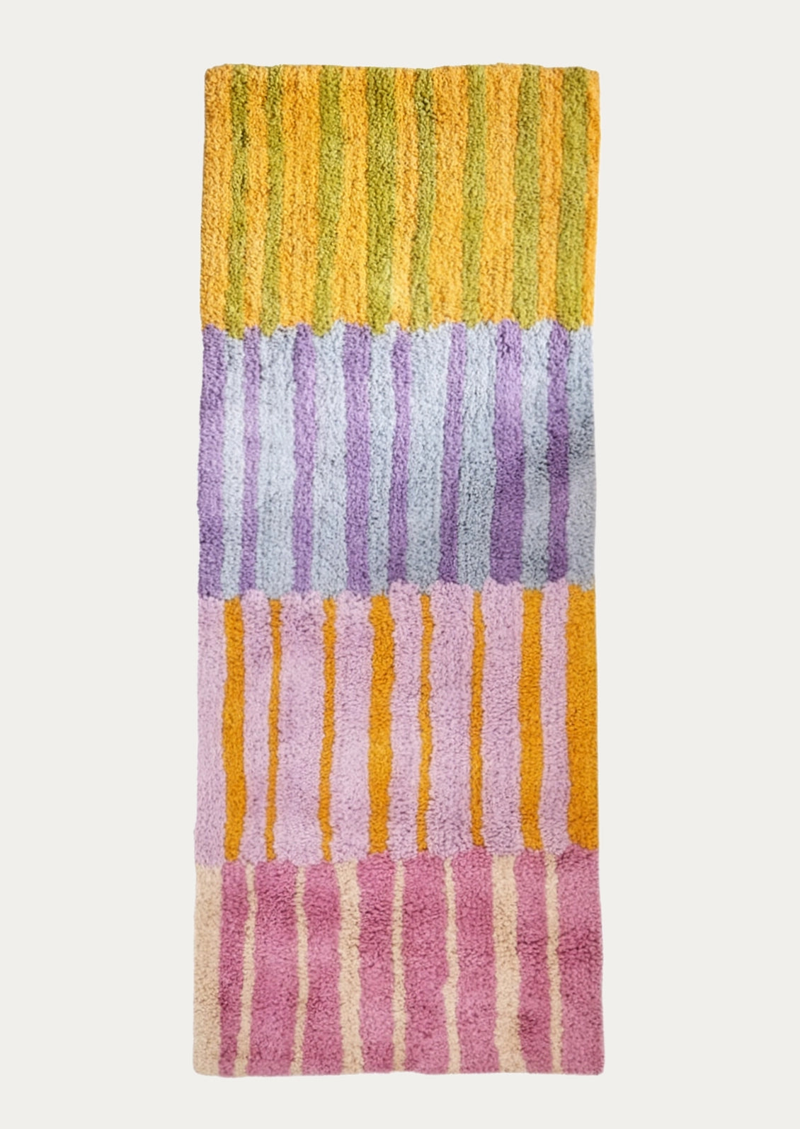 A rectangular plush bath mat with vertical striped blocks in yellow, lavender, pink, and cream, shown flat against a light gray background.