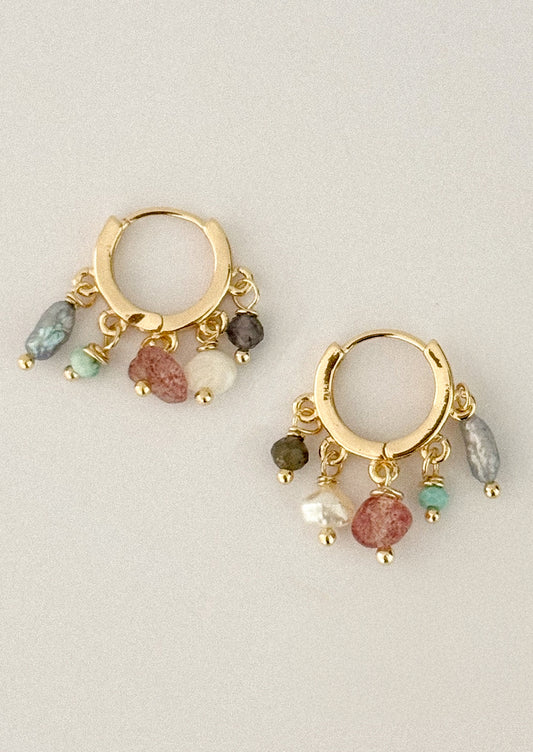 A pair of gold huggie hoop earrings with dangling multicolor gemstone charms, displayed on a neutral light gray background.