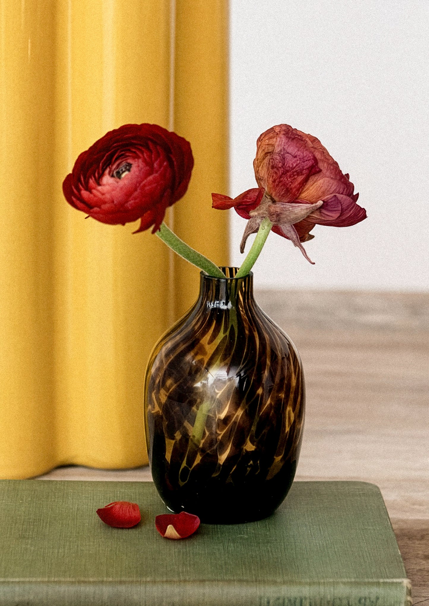 Tortoiseshell vase with wilted red flowers on a green surface against a yellow curtain.