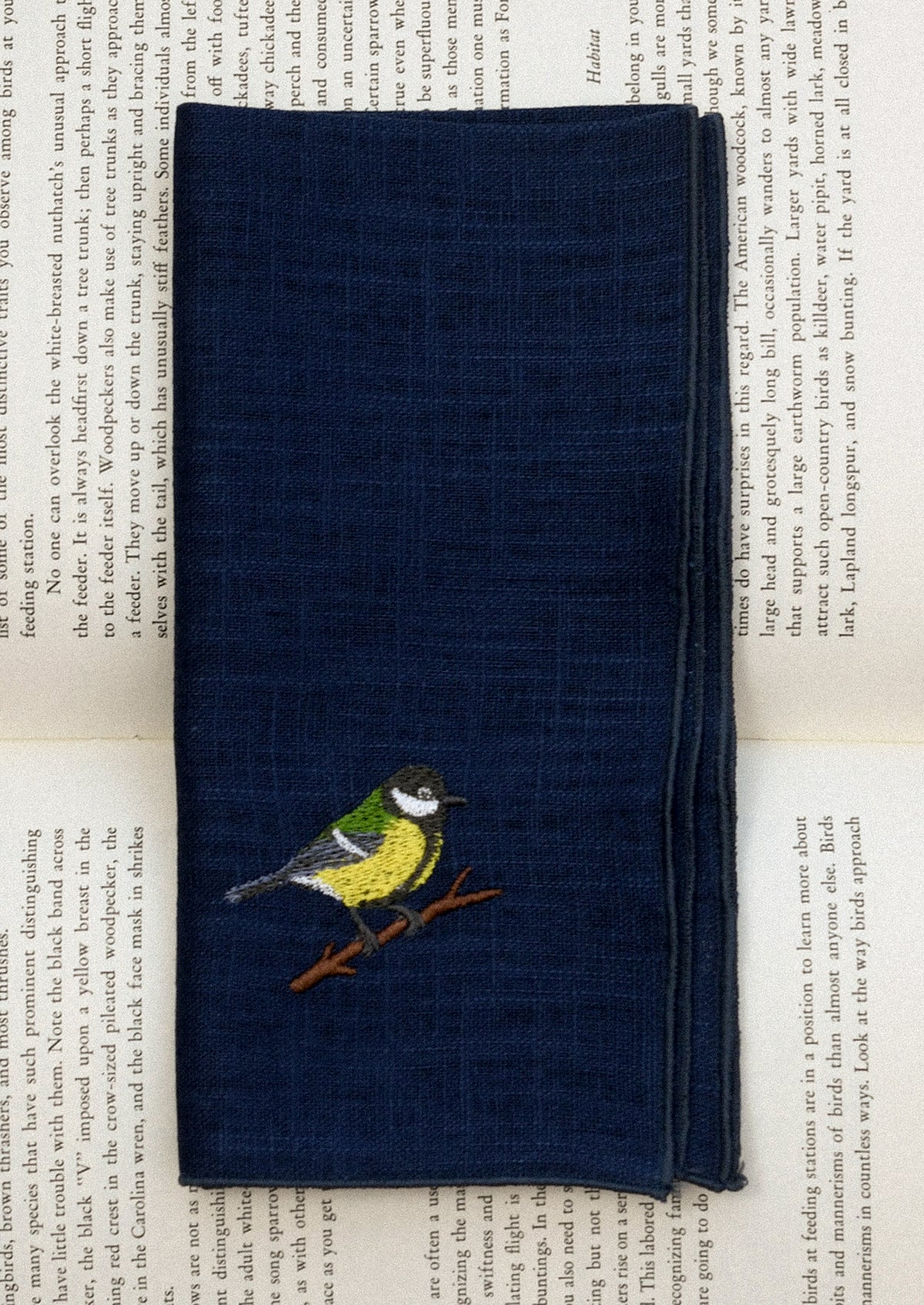 A navy blue linen napkin with tonal serged edges, and an embroidered warbler at corner.