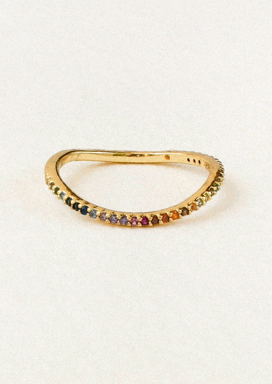 A gold band ring featuring a row of small, multicolored gemstones set along the front, photographed on a light off-white background.