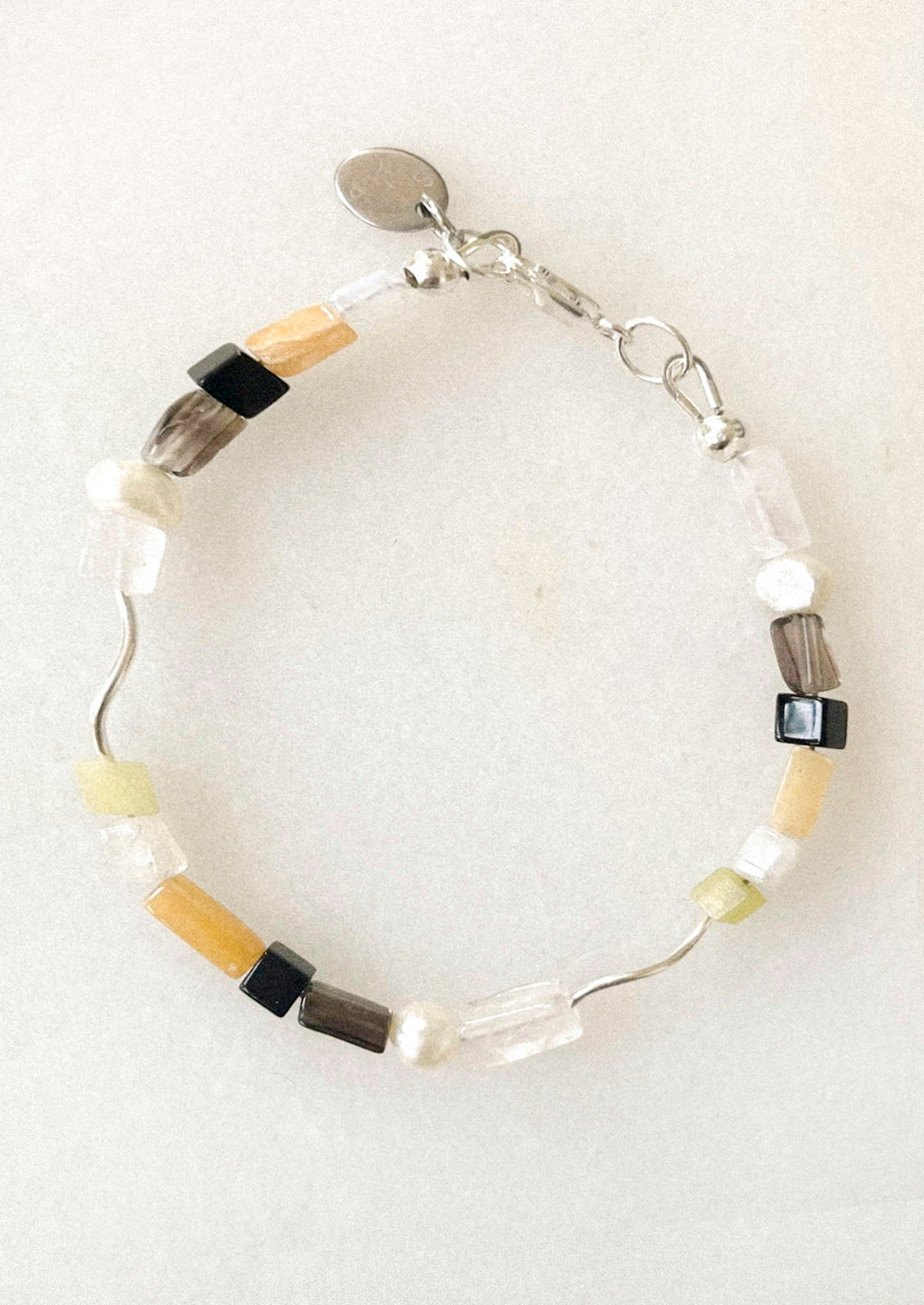 A bracelet with a wavy silver wire base, featuring clear faceted beads, light green beads, dark gray beads, and small black square beads, with a silver clasp and a small round tag.