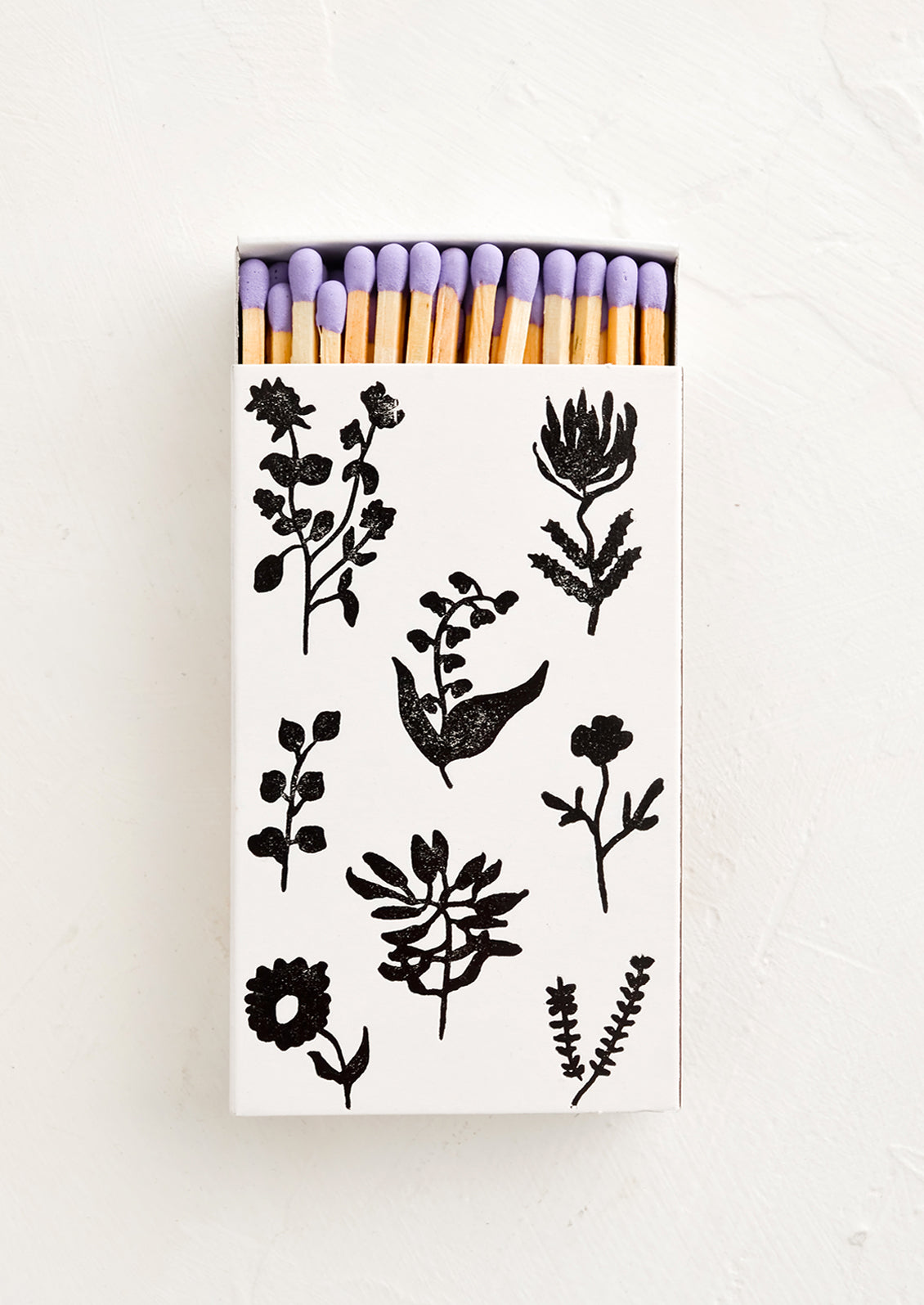 A matchbox in white with black botanical print, housing purple tipped matches.