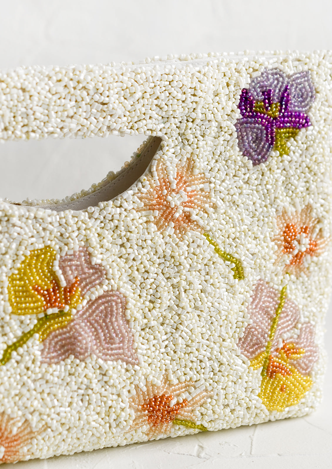 A white beaded east-west clutch with cutout handle and pastel floral pattern.