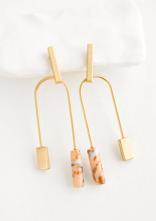 A pair of gold earrings with bar shaped top and arched bottom with peach gemstone on one side.