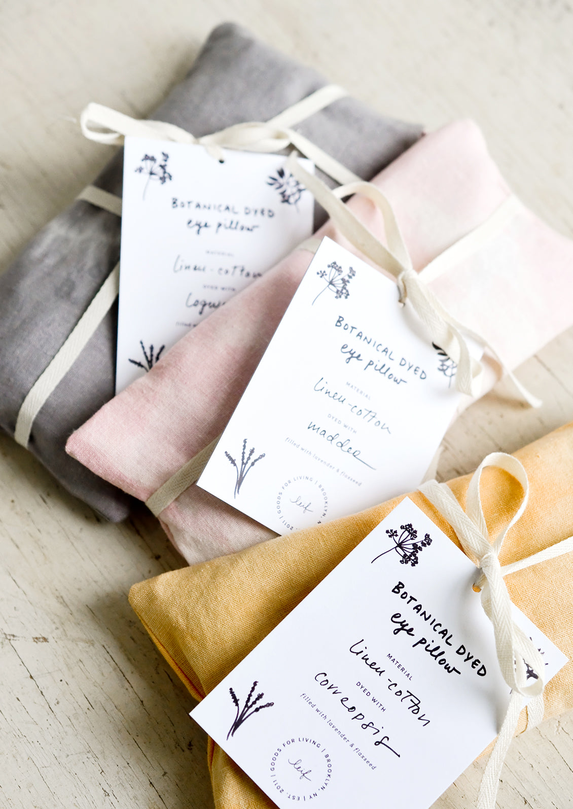 Three rectangular eye pillows in pink, orange and grey sitting on a tabletop.