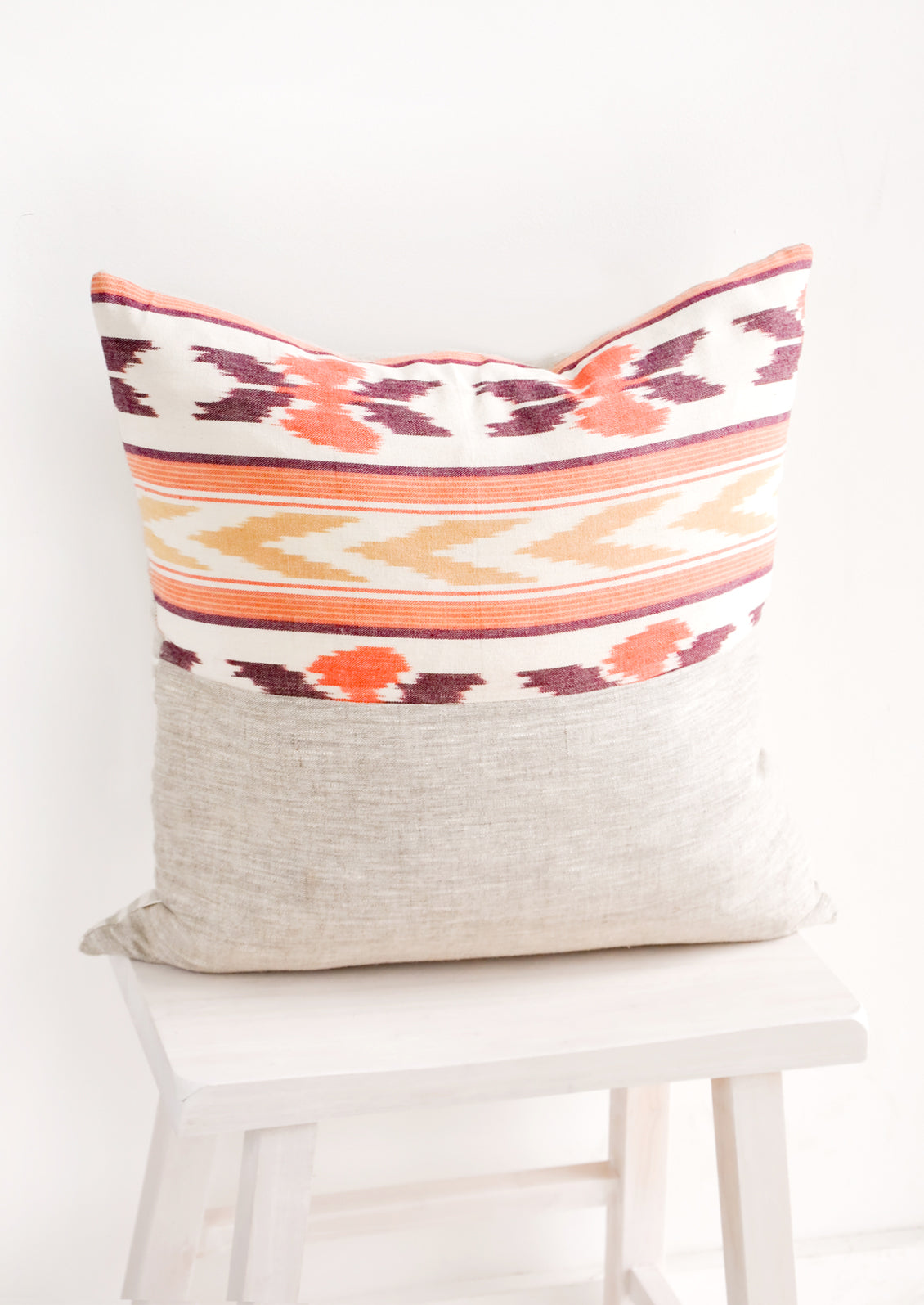 Square throw pillow in mixed fabrics. Top half is ikat print in orange and red, bottom half is plain linen.