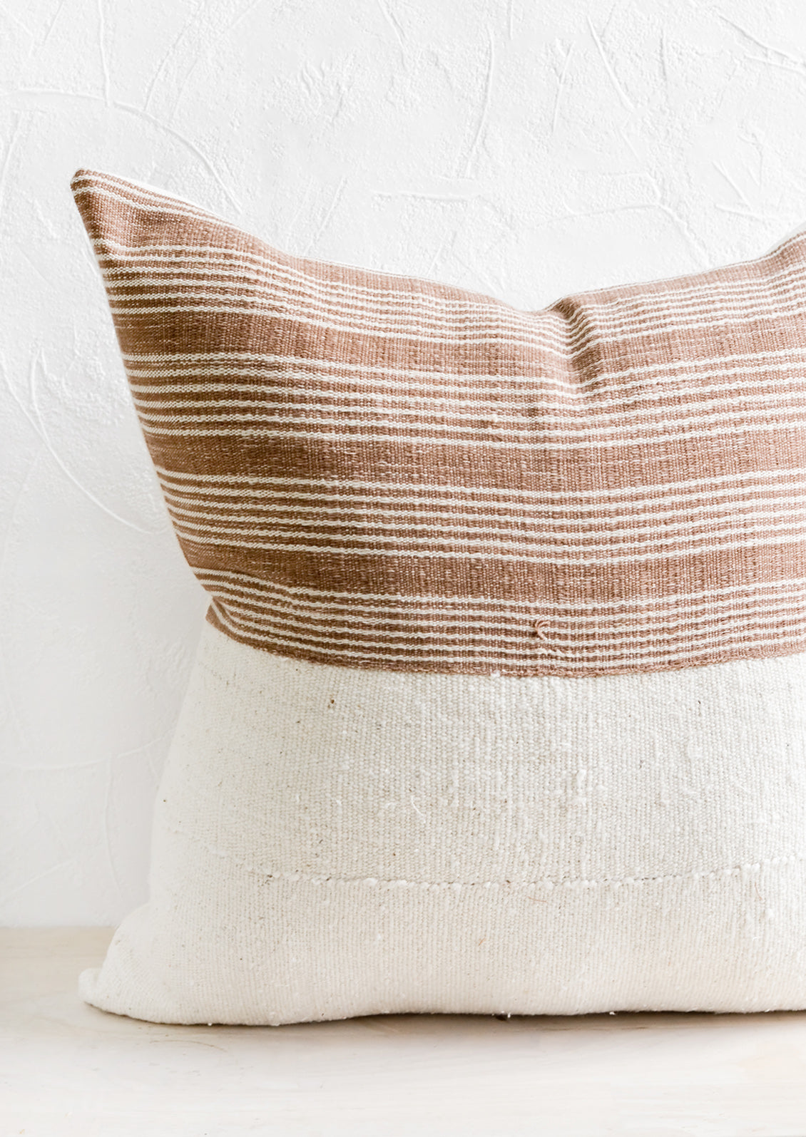 A throw pillow with top half in brown & ivory striped fabric and bottom half in natural mudcloth.