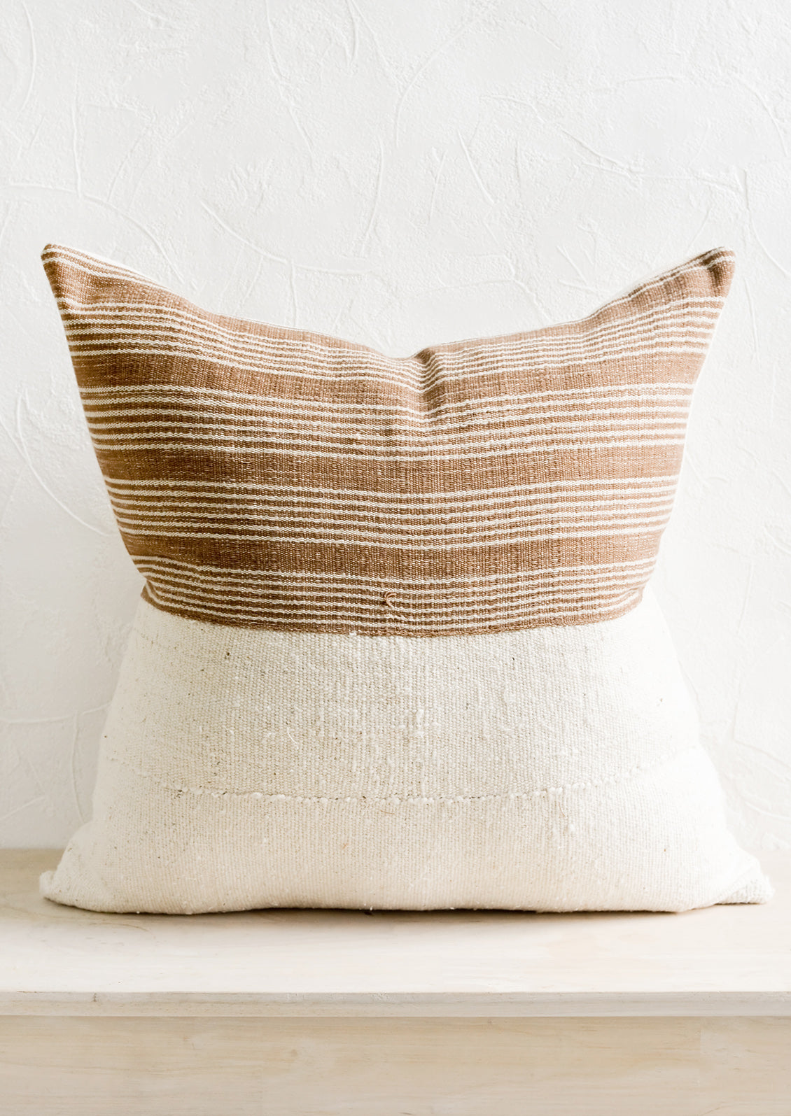A throw pillow with top half in brown & ivory striped fabric and bottom half in natural mudcloth.