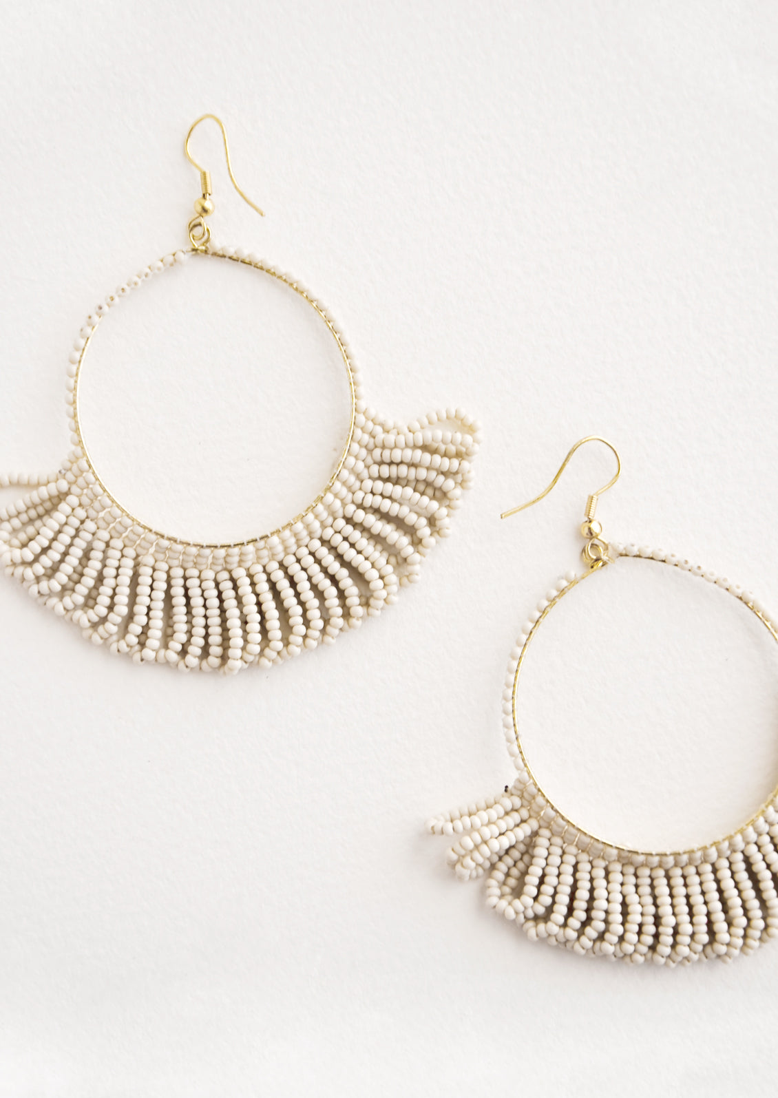Dangling hoop earrings featuring light grey beads and accented with hanging beaded, looped fringe.