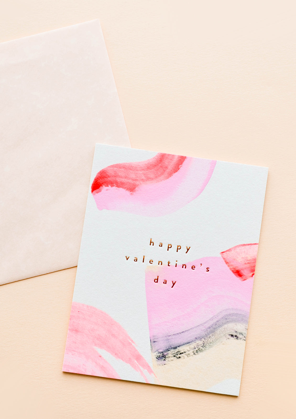 A beige envelope and white greeting card patterned with pink and red paint strokes reading "happy valentine's day" in gold foil.