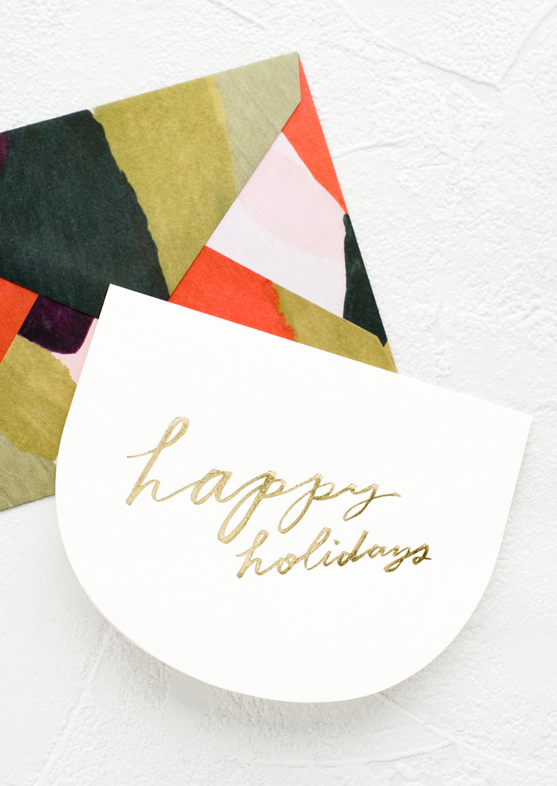 Greeting card in curved shape with gold script reading "Happy holidays", paired with green and red abstract envelope.