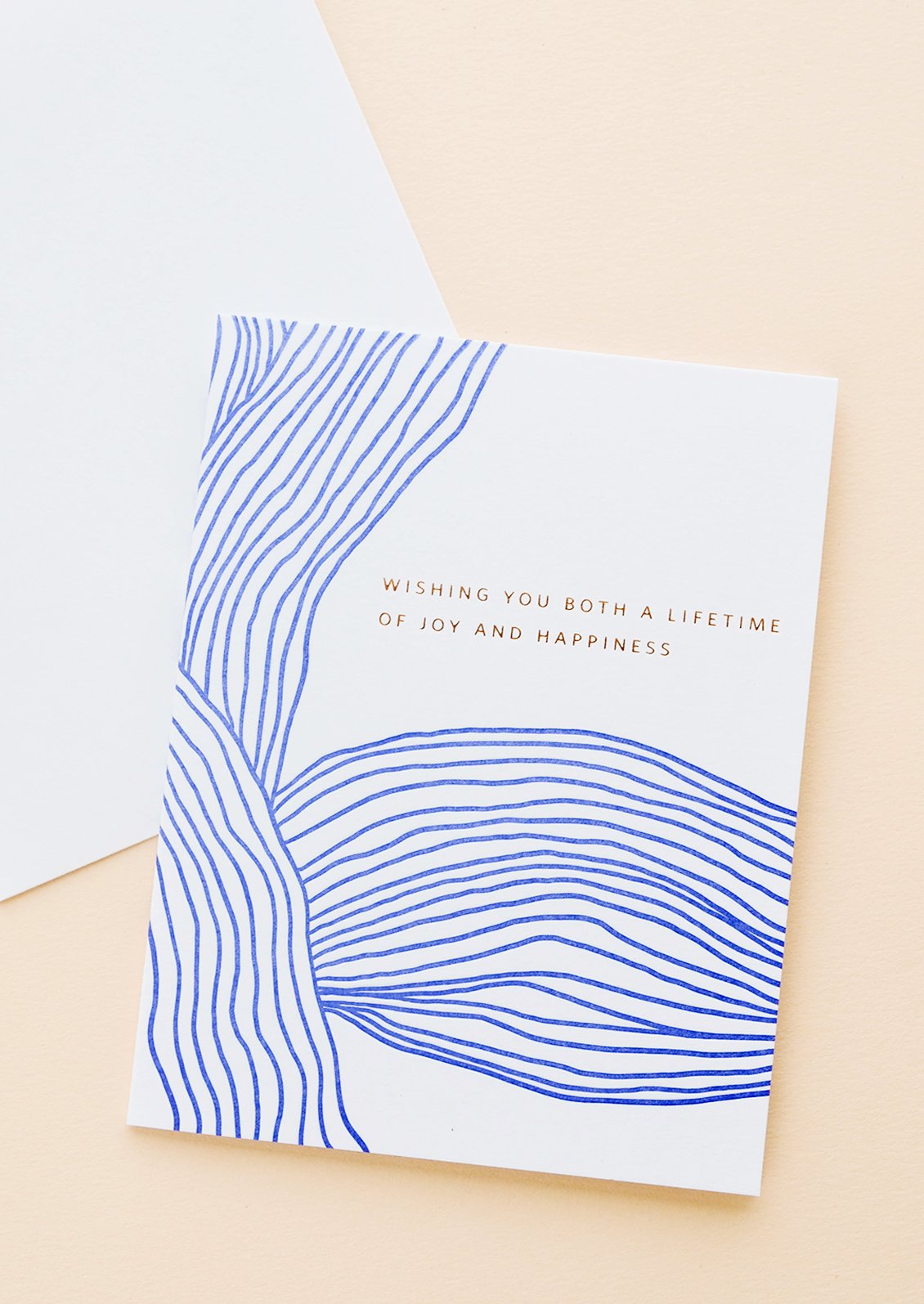 White greeting card with wavy cobalt blue line pattern and gold text reading "Wishing you both a lifetime of happiness"