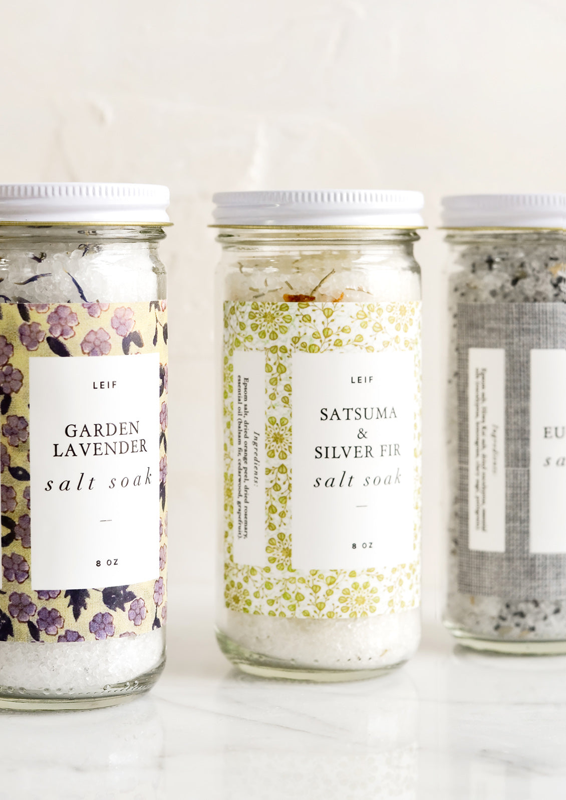 Three glass jars with decorative labels containing bath salt soaks.